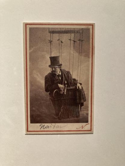 Photography of a man wearing a top hat sat in a tiny basket in the sky