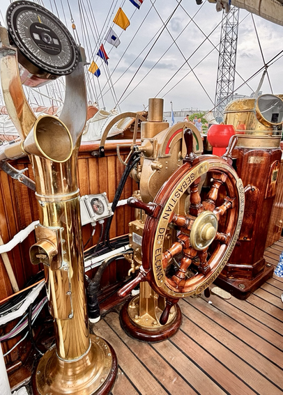 varnished wooden ship’s wheel, engraved with “Sebastián de Elcano,” identifying the Spanish Navy’s training ship. Surrounding the wheel is polished brass engine-order telegraph equipment, navigation instruments.