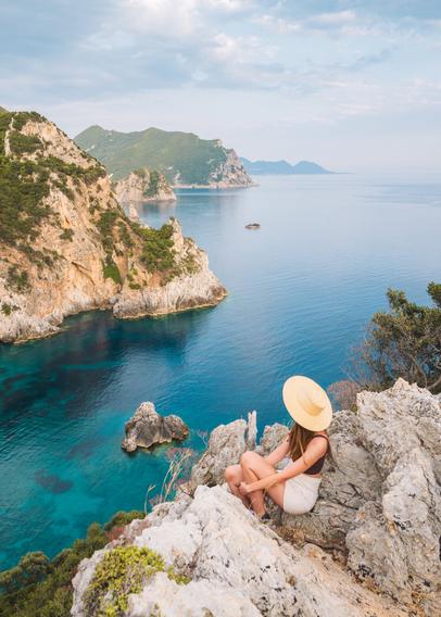 A girl wearing a hat is sitting on the edge of a rock, looking on a bay dotted with hills covered in greenery and trees. The sky has clouds during sunset. The water is crystal clear and blue.