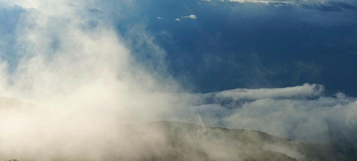 Sich auflösende Wolkenfetzen vor sehr dunklem Himmel über einem kaum erkennbaren Bergrücken.