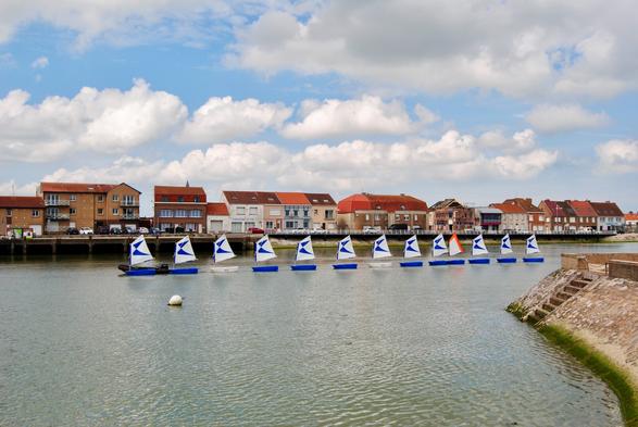 A view of a calm waterway lined with buildings featuring red rooftops. Several small sailboats with white sails and blue accents are seen sailing in a row, while fluffy clouds fill the sky.