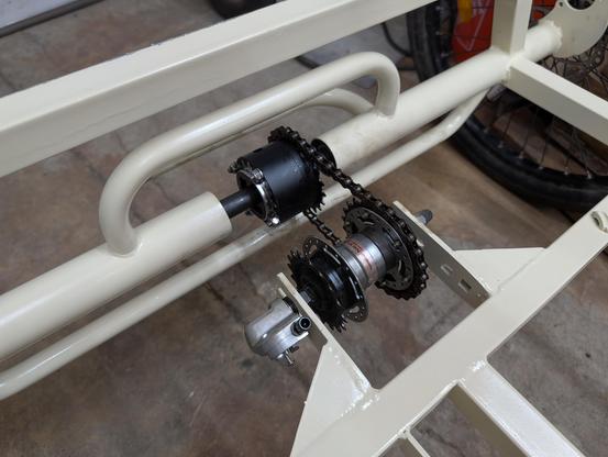 The center of the rear axle of a very large cargo trike. A short section of chain from a geared bike huh mounted in the frame runs to a stout cylinder on the axle, anodized black.