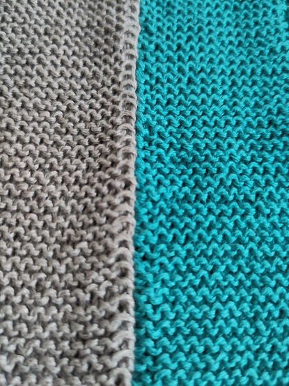 Zu sehen ist eine gestrickte Handarbeit, die nur aus rechten Maschen besteht. Auf der linken Seite ist graue Wolle erkennbar, auf der rechten Seite des Strickstücks ist die Wolle türkisfarben.