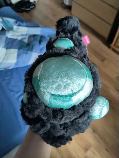 A top down view on the back of the plushie crystal lizard. It's body is roughly teardrop shaped, with the thick end being its head.
It has a total of 4 gems on its body. One very large in the center of its back and 3 differently sized ones arranged in a triangle around the center gem.