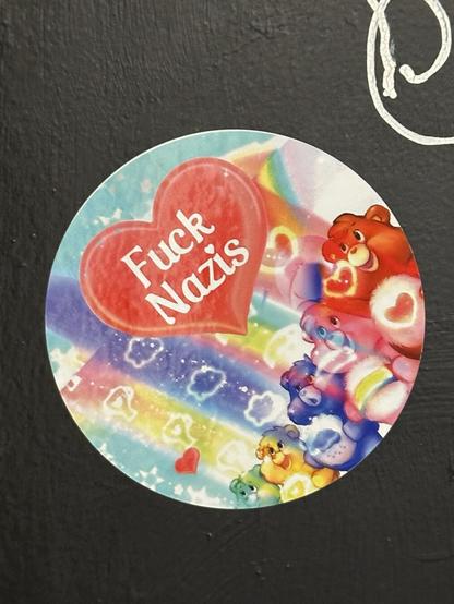 a round sticker with some Care Bears-style characters, rainbows, and a large red heart with the text “Fuck Nazis”