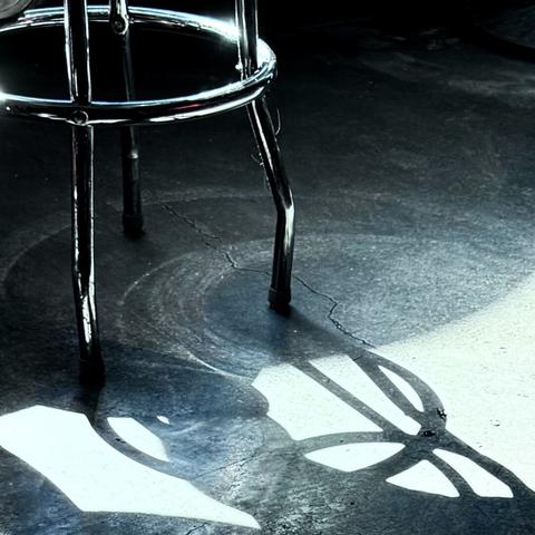 Chrome stool casting a shadow on a concrete floor. The photo appears to be in black and white, but there are small hints that it's not, such as a small orange reflection on the stool