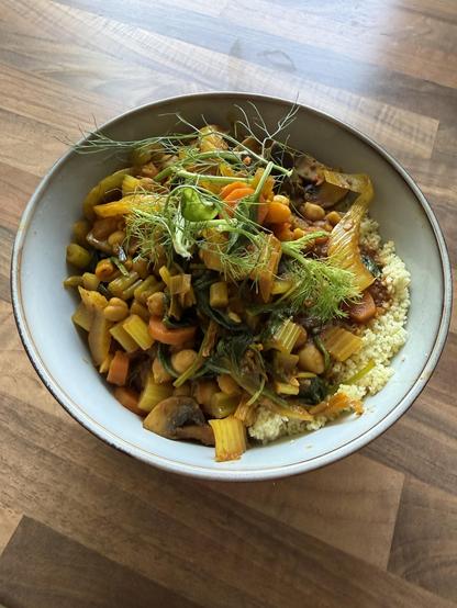 Eine Schüssel mit buntem gemischtem Gemüse, darunter Karotten, Sellerie und Gemüse, serviert auf einem Bett aus Couscous. Frische Kräuter garnieren das Gericht. Die Schüssel wird auf eine Holzoberfläche gestellt.