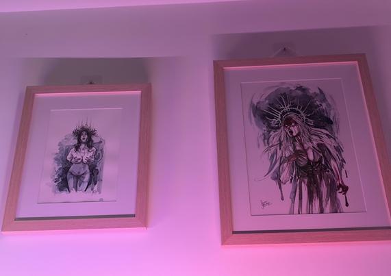 A pair of paintings hanging on a white wall. One painting is a nude Saint Lucia with her hands over her breasts, looking directly at the viewer. The other painting is an angry woman covered with blood roaring.