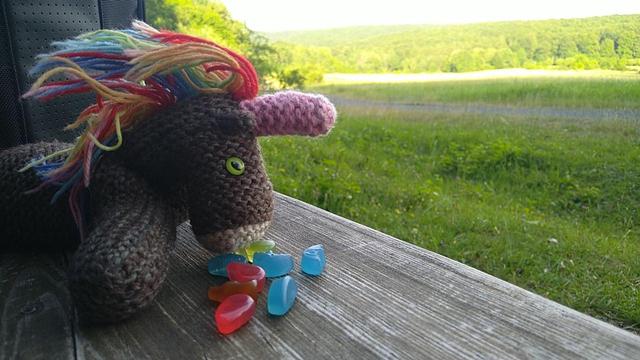 Max, das bunte Strickhorn, mit Fruchtgummi auf einer Holzbank im Grünen.