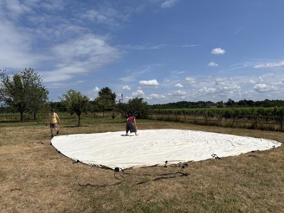 Une grande toile de tente blanche étalée dans un pré, deux personnes en train de la nettoyer. Ciel bleu avec des nuages moutonnants.