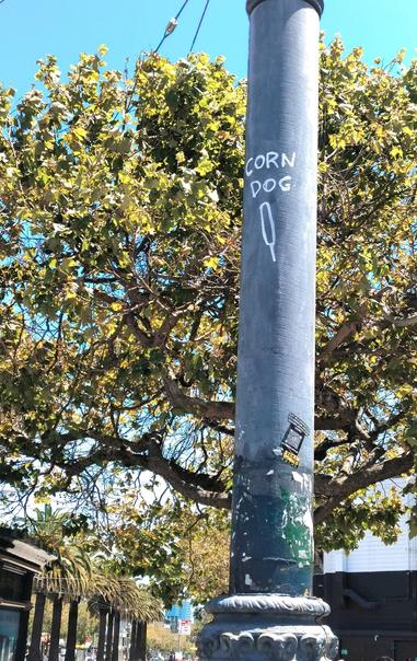 Street lamp pole with a very simple line drawing of a corn dog labeled with the text "corn dog".

You can't really tell from the photo but this is about eight feet (two and half meters) off the ground. Someone climbed the pole to draw this