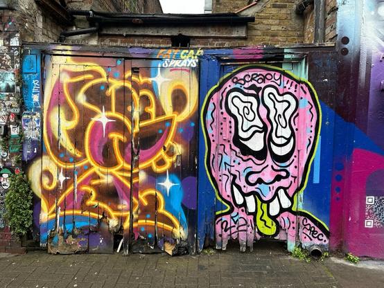 a wooden wall or fence in a laneway, thoroughly covered with colourful aerosol art. On the left is what looks like a Mickey Mouse-style cartoon character composed of glowing yellow lines over a coloured background, and on the right, a wobbly melting face in an underground-comic style