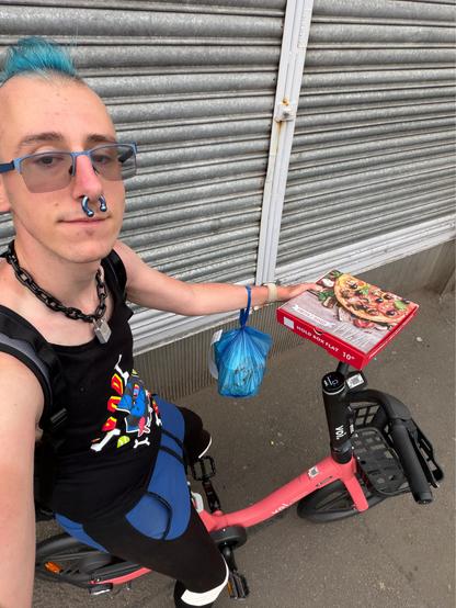 wide-angle selfie where I’m sitting on one of the Voi rental e-bikes, with a blue plastic bag containing food hanging from my left arm, and a 10” pizza box in my left hand