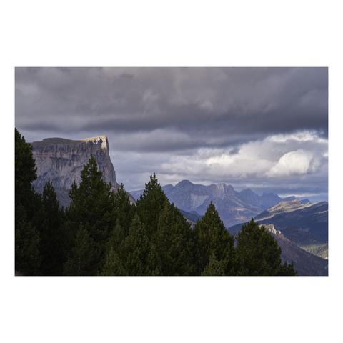 Photo d'un paysage de montagne.
Le paysage est dominé sur la gauche par une montagne aux flancs escarpés, mais au sommet plat couvert d'une prairie. Un rayon de soleil éclaire juste la point qui domine la falaise à droite. La montagne se dresse au-dessus d'une forêt de pins au premier plan. 
Au dernier plan, d'autres montagnes meublent le paysage, quelques rayons de soleil éclairent des zones aléatoires. Le ciel est très nuageux.
-
Photo of a mountain landscape.
The landscape is dominated on the left by a mountain with steep slopes, but with a flat summit covered in a meadow. A ray of sunlight illuminates just the peak that rises above the cliff on the right. The mountain rises above a pine forest in the foreground. 
In the background, other mountains dot the landscape, with a few rays of sunlight illuminating random patches. The sky is very cloudy.