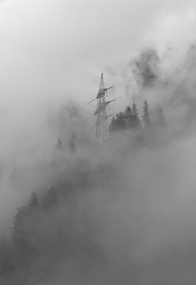 A high voltage transmission line pylon surrounded by suddenly formed thick clouds, rising above the trees partially obscured in the mountainside.
