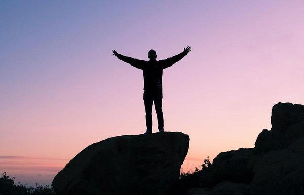 a silhouette of a person standing on a large rock with arms outstretched against a pink and blue twilight sky