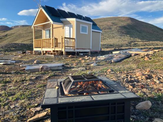 A small fire in a small fire pit ringed with a slate border. In the background is our cabin and green hills. There is a bright blue sky dotted with white clouds.