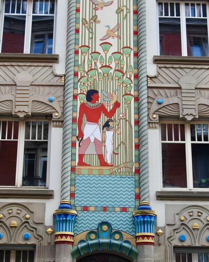 Artwork depicting a figure in ancient Egyptian attire holding a bird, accompanied by a smaller figure. The design features stylized plants and birds, rendered in vibrant colors against a decorative background, showcasing an architectural element.
