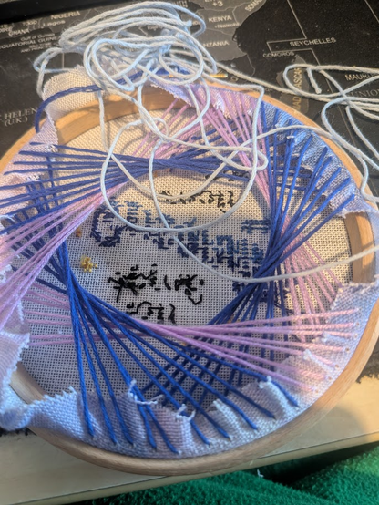 The back of a cross stitch in a small wooden hoop. The back of the pattern is covered in long thread pulled tight. The base layer of the threads had three points of contact, initially stitching a triangle, this has continued round the back of the pattern to cover the back and tighten the fabric. It looks like a tangle of threads.