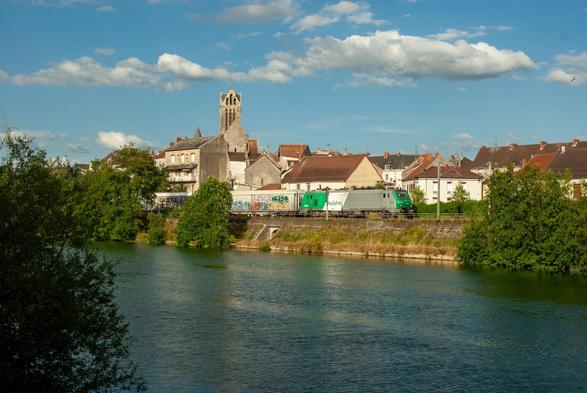 A Hexafret "Prima" locomotive hauls a train of covered freight waggons along the Marne, past the village of Dormans with its impressive church behind the train.