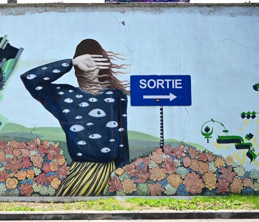 A mural depicting a figure with long hair, wearing a sweater adorned with multiple eyes, shielding their face with one hand. In the background, there's a blue sign reading "SORTIE" with an arrow pointing to the right, next to colorful flowers.
