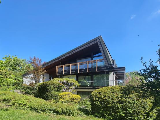 The image shows a house with a steeply pitched roof, set within a well-maintained garden and framed by mature vegetation under a blue sky. The building features large windows, dark roof cladding, and a wooden balcony, creating a strong architectural presence, while the surrounding greenery lends the scene a calm and balanced character.