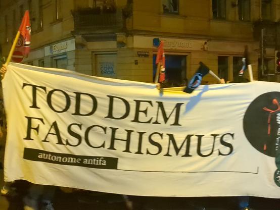 Großes weißes Banner mit schwarzer Aufschrift wird von mehreren Personen bei Dunkelheit auf einer Straße getragen. Auf dem Banner steht in großen Buchstaben: 'TOD DEM FASCHISMUS', darunter in kleinerer Schrift: 'autonome antifa'. Links und rechts vom Banner sind rote und schwarze Fahnen mit Symbolen zu sehen. Im Hintergrund beleuchtete Fassade eines mehrstöckigen Gebäudes.