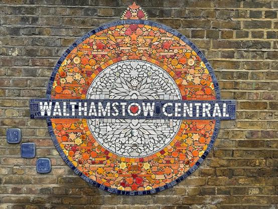 a wall with a large mosaic of patterned/textured orange, white and blue ceramic tiles, forming a roundel with the text WALTHAMSTOW CENTRAL on a bar across the centre