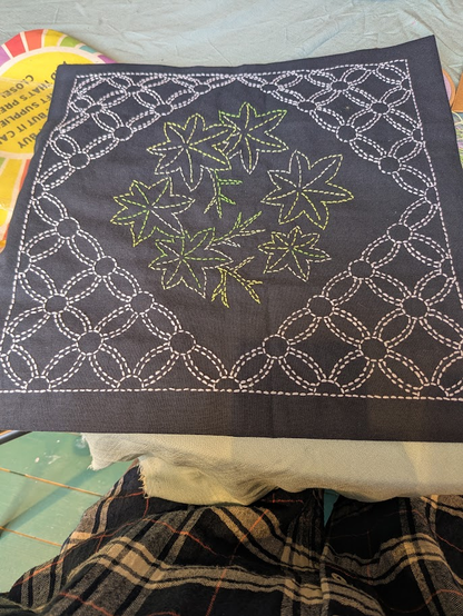 Navy fabric, square shape. Each corner is decorated with white stitching, like flowers, the centre is a cluster of leaves which are stitching in a variegated green thread.