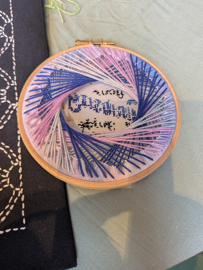 The back of a cross stitch hoop, finished in a "string art" style manner, 3 different colours of threads over lapping each other, making circle in the middle.