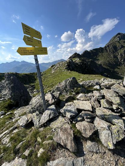 A signpost with directional arrows and destinations against an alpine backdrop. The landscape features rocky terrain and patches of grass under a blue sky with cumulus clouds. The sign indicates hiking routes and gives time estimates.
