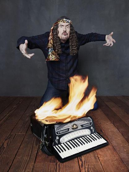 "Weird Al" Yankovic kneels with his arms outstretched, with a burning accordion beneath him.