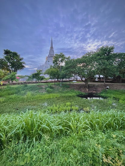 Up close to us are grassy green weeds waving, rising from a klong/canal/moat that goes around the temple. We see the darkness of water, crowded with many types of watercress, lilies and grasses, a round portal extends over the water, the perfect place to pop through and visit anytime you wish. Beyond we see trees and red brick ruins and then the great white temple, Wat PhuKhao Thong rises majestically into the cloudy Grey skies of a storm approaching. Serene.