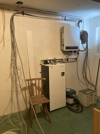 Keller. Weiße Wand. Ein turmartiges Gerät steht auf dem Boden. Ein weißer Kasten hängt an der Wand. An der Wand sind Kabelkanäle festgeschraubt. Viele Kabel hängen noch lose herum.