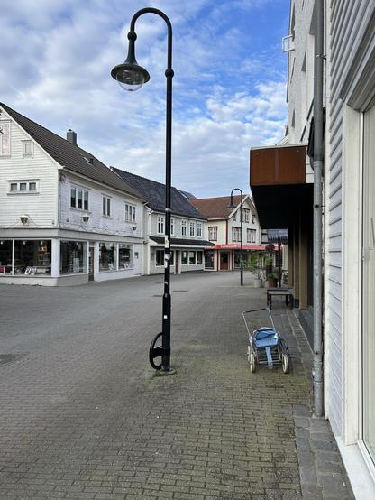 A quiet street scene featuring a lamp post and a blue baby stroller parked on the sidewalk. White wooden buildings with colorful details line the street under a partly cloudy sky.