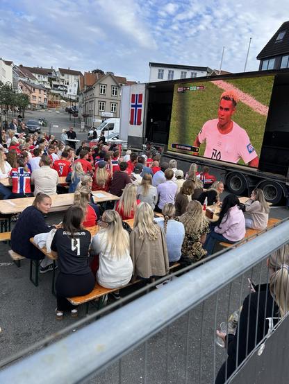A large outdoor crowd is gathered on benches watching a football match displayed on a big screen mounted on a truck. Many attendees are wearing red jerseys, and a flag of Norway is visible in the background. The atmosphere appears lively, with a mix of men