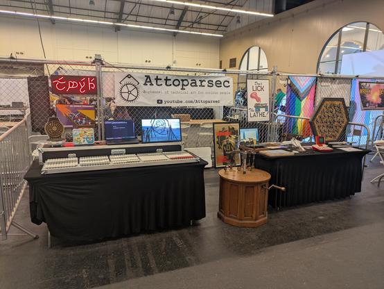 The Attoparsec booth at Open Sauce 2025. A double-wide booth with 2 tables covered in past projects. The chainlink fence divider at the back is hung with shop posters and a neon sign in Elvish. A kinetic bird sculpture sits on the ground in front of the tables.