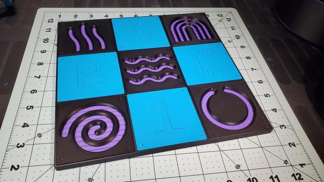 A 9x9 modular tacile sensory board, fitted with black stencils, lavender textures, and cyan solid plates.