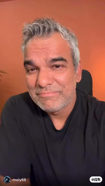 grey haired nan with brown skin and eyes in black shirt in front of an orange wall.