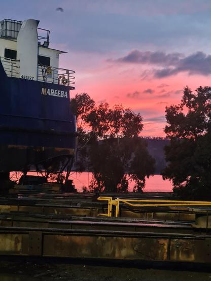 Aft end of steel ship named "Mareeba" sillhoutted with trees agains a pink dawn sky which us reflected in the Derwent River. Steel beams and timbers in the foreground.