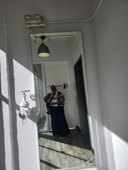 Mirror selfie in a tiny elevator corridor of a Black woman feeling cute wearing lots of fabric: Wide legged denim trousers with a button+zipper fly; graphic dress worn as a shirt; heavy gold chain; camo LA Dodgers ball cap. Animal print pointy toed flats.
