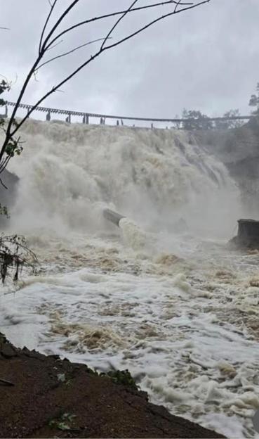 A photo of a raging torrent of water, a waterfall over an embankment. A pipe sticks out of the middle of the torrent as shooting water. Above the torrent train tracks can be seen, partially suspended, partially on piles?