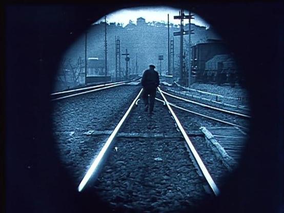 A circular frame of a man in silhouette walking down the center of some very shiny train tracks. The camera is also positioned in the center so the man's body obscures the two parallel extending to infinity. In the far distance is a hill with some buildings and trees on it. You can wonder if there's a tunnel for the tracks. The image monochrome, but tinted blue, a common way of marking outdoor daytime shots in silent films.