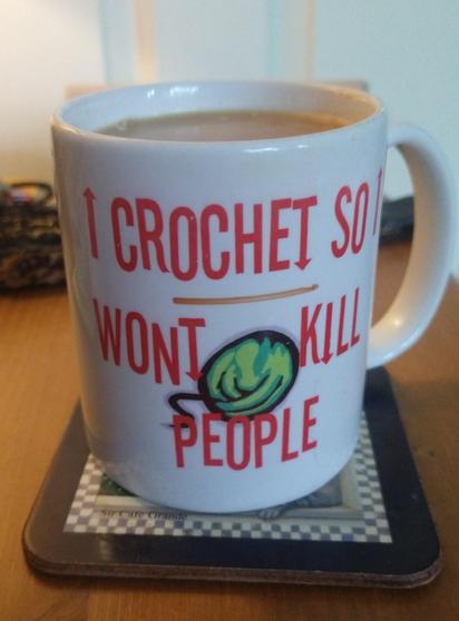 Mug of tea on a bedside cabinet with red text saying I crochet so I won't kill people it has a horizontal crochet hook in the middle above a ball of green yarn