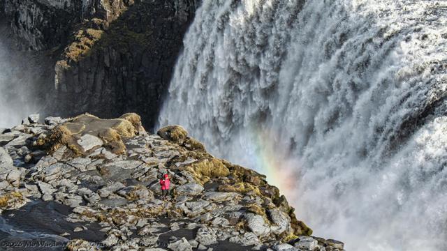 A colour photo of a waterfall taken from above. The cascade is only partially seen, stretching from centre top to bottom right of the shot and dropping into an unseen canyon far below. Some spray is rising on the far left. The water is grey with sediment and foaming white from before the fairly straight crest line. At the top left is the opposite wall of the canyon, craggy brown rock mostly in the shadow of the waterfall. Directly below the high viewpoint is an outcrop, grey rock and dark mud with some scrubby plant growth. Walking across it is a photographer in a red coat and hat with a tripod over his shoulder. At the edge of the outcrop, the dropping water is creating spray and a small arc of rainbow is visible.