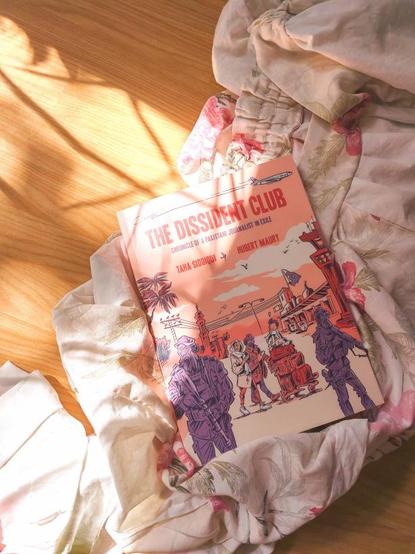 photo of Taha Siddiqui and Hubert Maury's comic book The Dissident Club lying on a pile of similarly colored fabric of white-ish  green and cream with red and pink flowers, all on an imitation wood floor with perfectly aligned ray of light gently caressing them