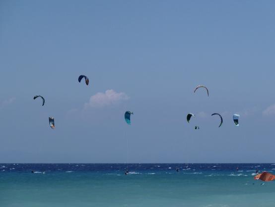 A clear blue sky above a vibrant sea features several colorful kitesurfing kites in the air, with kitesurfers visible on the water below.