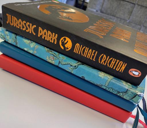 A stack of three hardcover notebooks and a paperback copy of Jurassic Park by Michael Crichton.