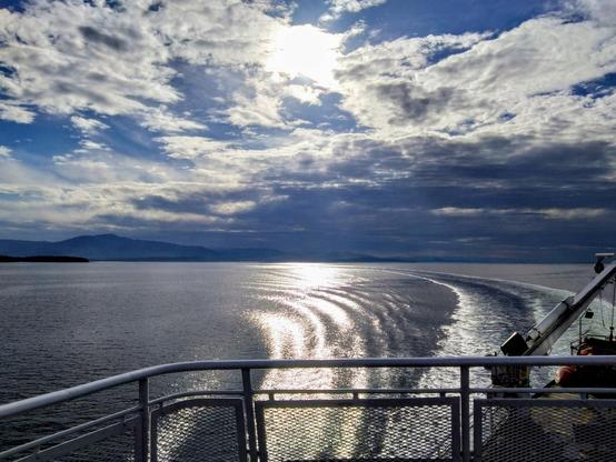 Sicht vom Deck einer Fähre. Ein Tel des Geländers ist zu sehen, ausserdem ein Rettungsboot und ein kleiner Kranarm. Da der Blick gegen die Fahrtrichtung geht, sieht man die Fahrspuren im Meer. Im Hintergrund ist schwach eine hügelige Landmasse erkennbar. Am Himmel sind Wolken zu sehen, die Sonne ist aber erkennbar.