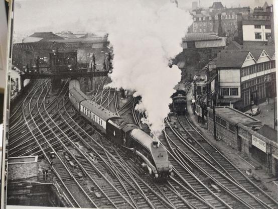is black and white photo from above looking into railway station throat with loads of points and crossovers. Is BR liveried A4 pulling train out of station under big plume of exhaust with 3 coaches visible behind.
